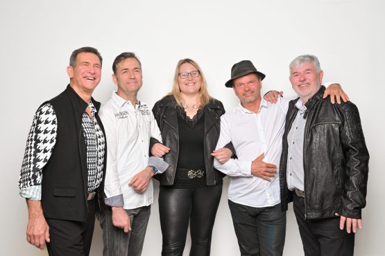 Von links: Bert, Torsten, Leni, Mirko und Uwe stehen vor einem weißen Hintergrund und lächeln in die Kamera. Bert trägt ein schwarz-weiß gemustertes Hemd mit einer schwarzen Weste, Torsten ein weißes Hemd mit Schriftzügen, Leni eine schwarze Lederjacke und schwarze Lederhose, Mirko ein weißes Hemd und einen schwarzen Hut, und Uwe eine schwarze Lederjacke mit einem gemusterten Hemd darunter.