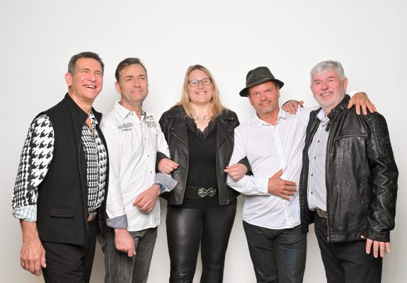 Von links: Bert, Torsten, Leni, Mirko und Uwe stehen vor einem weißen Hintergrund und lächeln in die Kamera. Bert trägt ein schwarz-weiß gemustertes Hemd mit einer schwarzen Weste, Torsten ein weißes Hemd mit Schriftzügen, Leni eine schwarze Lederjacke und schwarze Lederhose, Mirko ein weißes Hemd und einen schwarzen Hut, und Uwe eine schwarze Lederjacke mit einem gemusterten Hemd darunter.