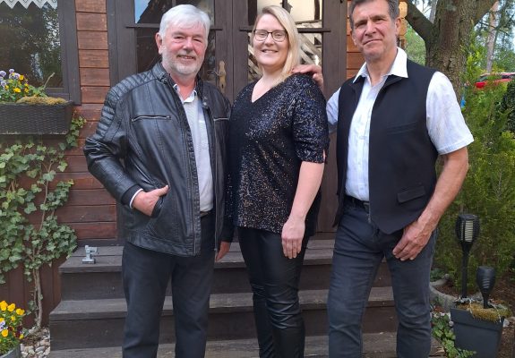 Uwe, Leni und Bert stehen lächelnd vor einer kleinen Holzhütte. Uwe trägt eine schwarze Lederjacke, Leni ein schwarzes Paillettenshirt und Bert ein weißes Hemd mit schwarzer Weste. Die Szene ist von einem Garten mit Blumen und Bäumen umgeben.