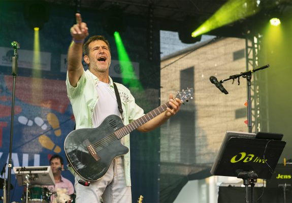 Bert, Gitarrist und Sänger, steht auf der Bühne und singt leidenschaftlich, während er eine schwarze Gitarre spielt. Er trägt ein helles Hemd über einem weißen T-Shirt und hebt die Hand, um das Publikum zu animieren. Im Hintergrund sind Scheinwerfer und ein Schlagzeuger zu erkennen.