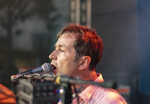 Torsten, der Schlagzeuger der Band, singt in ein Mikrofon. Er trägt ein helles Hemd und ist von Bühnenlicht beleuchtet, während er sich auf seinen Gesang konzentriert. Im Hintergrund sind verschwommene Bühnenelemente zu erkennen.