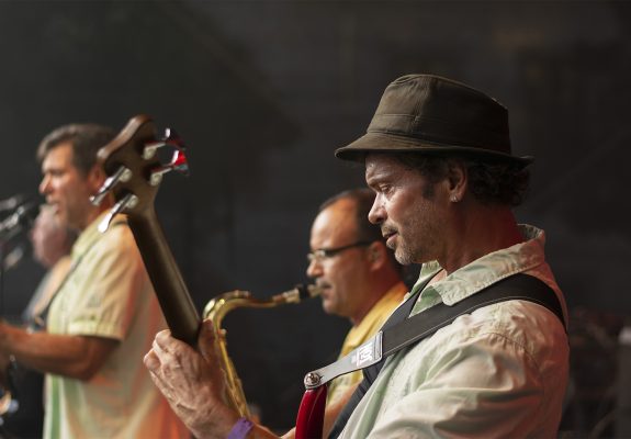 Mirko, ein Musiker mit Hut, spielt konzentriert Bass auf einer Bühne. Im Hintergrund sind unscharf weitere Bandmitglieder zu sehen, darunter ein Saxophonspieler und ein Gitarrist, die ebenfalls in die Musik vertieft sind.