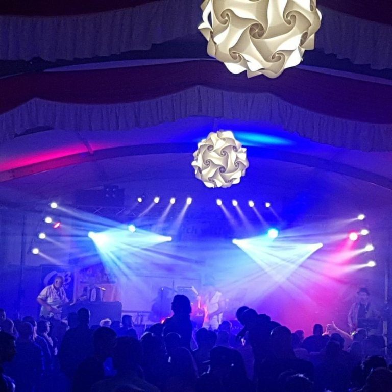 Auftritt der Band OBlive (Abkürzung für Oberlandbub’n) in einem Festzelt. Die Bühne ist in buntem Licht mit blauen, roten und weißen Scheinwerfern beleuchtet. Im Vordergrund tanzt und feiert das Publikum, während im Hintergrund die Band spielt. Dekorative Lampen hängen von der Zeltdecke.