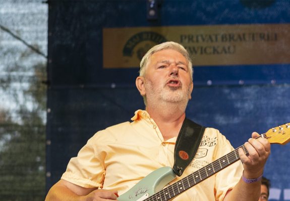 Uwe spielt auf einer Bühne eine mintgrüne E-Gitarre, während er singt. Er trägt ein hellgelbes Hemd und hellblaue Jeans. Im Hintergrund sind ein Keyboardspieler sowie Werbebanner sichtbar.