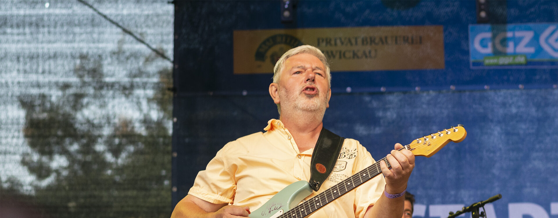 Uwe spielt auf einer Bühne eine mintgrüne E-Gitarre, während er singt. Er trägt ein hellgelbes Hemd und hellblaue Jeans. Im Hintergrund sind ein Keyboardspieler sowie Werbebanner sichtbar.