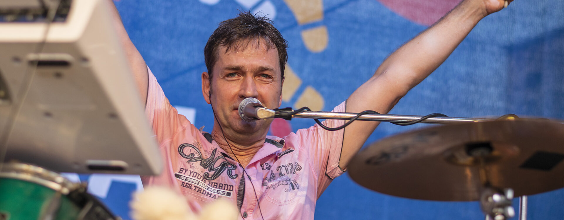 Torsten, der Schlagzeuger, hebt auf der Bühne mit einem Lächeln die Arme, während er in ein Mikrofon singt. Er trägt ein rosafarbenes Hemd und sitzt hinter seinem Schlagzeug. Im Hintergrund ist ein farbenfrohes Bühnenbanner sichtbar.