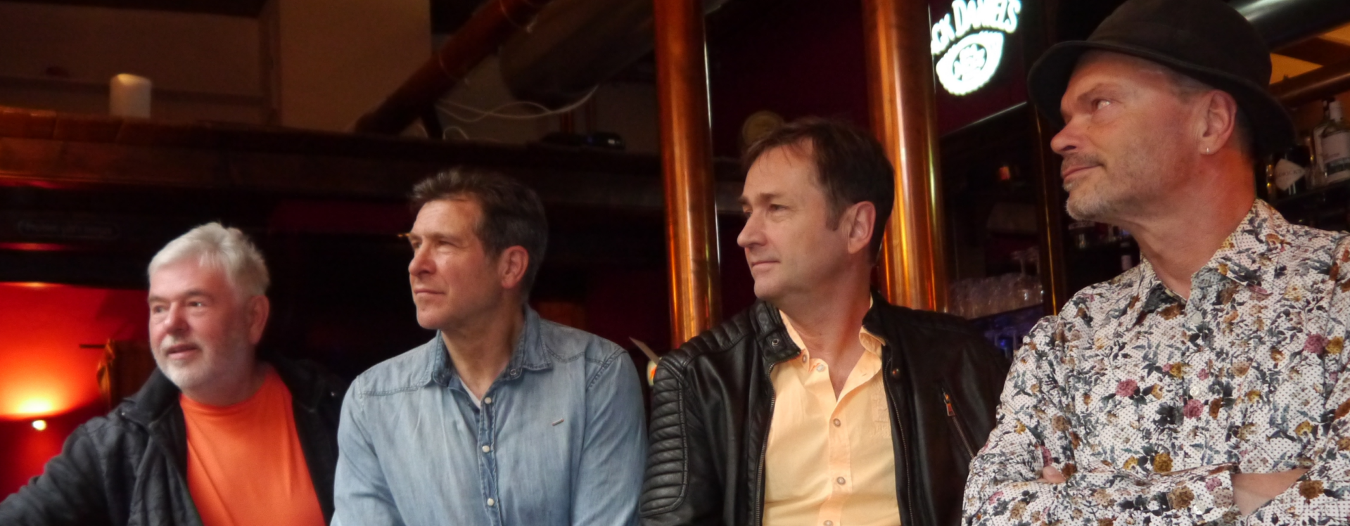 Uwe, Bert, Torsten und Mirko sitzen in einer Bar oder einem Restaurant und blicken gemeinsam in dieselbe Richtung. Uwe trägt ein orangefarbenes Shirt mit schwarzer Jacke, Bert ein hellblaues Hemd, Torsten ein gelbes Hemd mit schwarzer Lederjacke und Mirko ein gemustertes Hemd mit Hut. Die warme Beleuchtung und das rustikale Ambiente im Hintergrund schaffen eine gemütliche Atmosphäre.